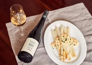 Weißwein neben Spargel mit Sauce auf weißem Teller, Holzuntergrund.