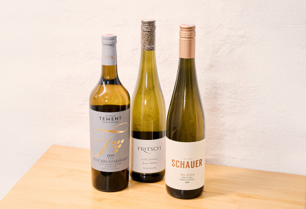 Drei Weißweinflaschen auf hellem Holztisch vor weißer Wand.