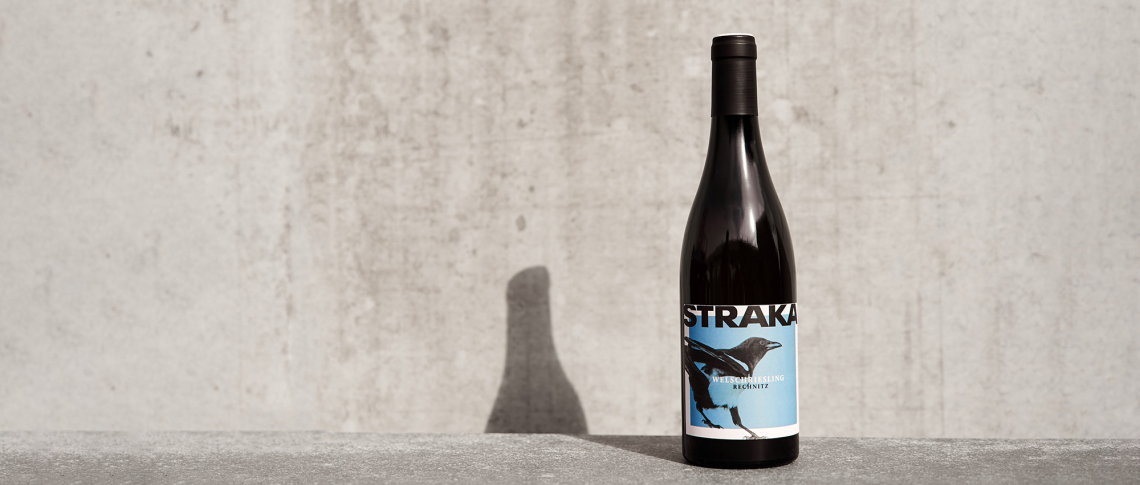 Dunkle Weinflasche mit blauem Etikett, vor grauem Betonhintergrund.