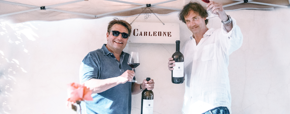 Zwei Männer halten Weinflaschen und Gläser unter einem Pavillon hoch.