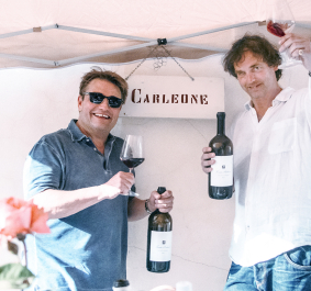 Zwei Männer halten Weinflaschen und Gläser unter einem Pavillon hoch.