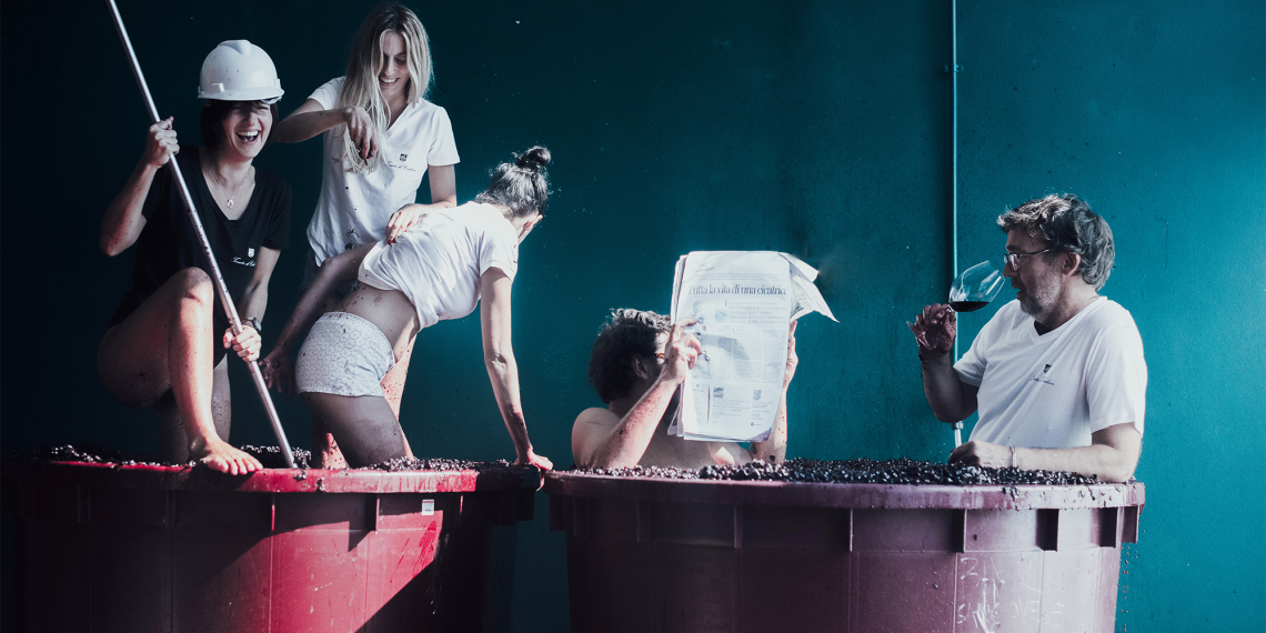 Gruppe von Menschen in großen Weinfässern, teils lesend, teils Wein trinkend.