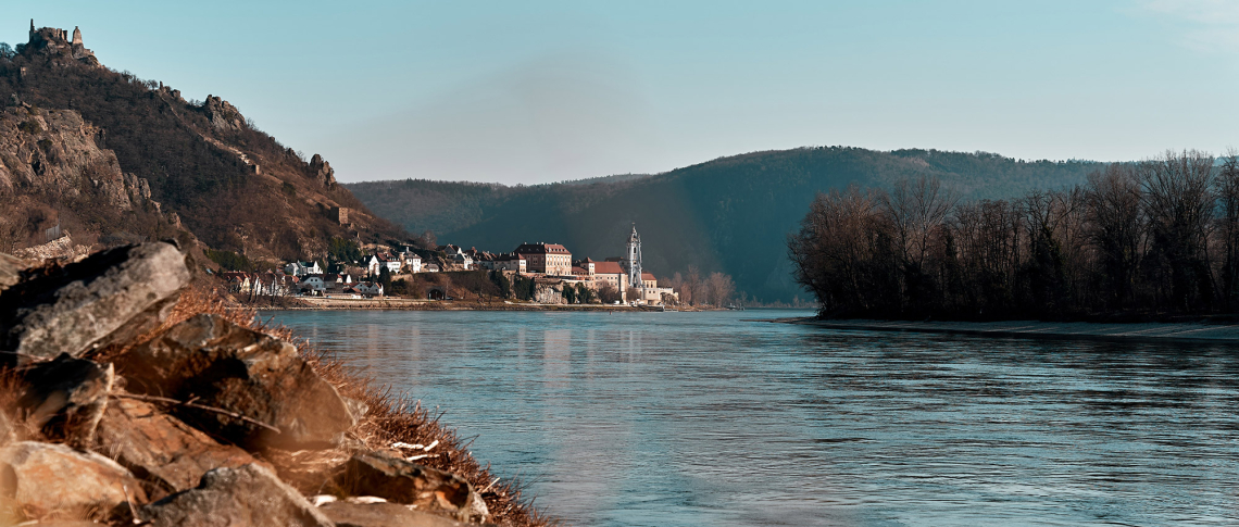 Flusslandschaft mit Dorf, Schloss und bewaldeten Hügeln unter klarem, blauem Himmel.