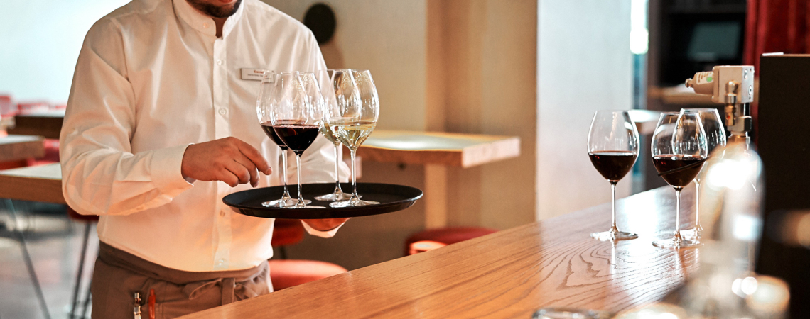 Kellner in weißem Hemd serviert Rot- und Weißweingläser auf Tablett in stilvoller Bar.