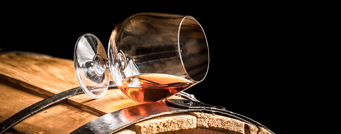 Weinglas mit Brandy auf Holzfass, vor schwarzem Hintergrund.