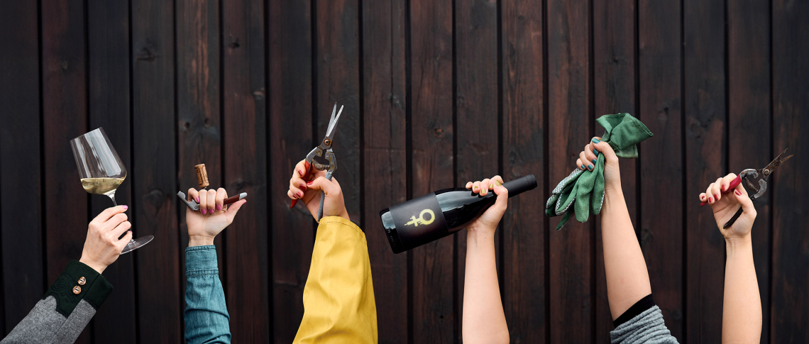 Fünf Hände halten Wein, Schokolade, Schere, Handschuhe vor dunkler Holzwand.