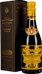 Eine elegante Flasche Balsamico-Essig mit einer schwarzen, glänzenden Oberfläche und einem gelben Etikett mit Goldverzierungen. Sie steht vor einer dunkelbraunen Kartonverpackung mit goldfarbener Schrift und Medaillon-Dekorationen.