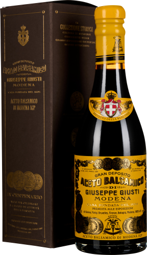 Eine elegante Flasche Balsamico-Essig mit einer schwarzen, glänzenden Oberfläche und einem gelben Etikett mit Goldverzierungen. Sie steht vor einer dunkelbraunen Kartonverpackung mit goldfarbener Schrift und Medaillon-Dekorationen.