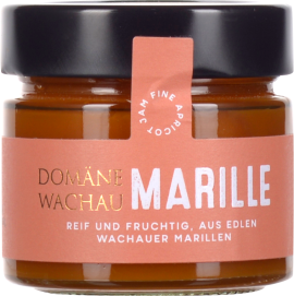 Ein Glas mit dunklem Schraubverschluss, gefüllt mit orangefarbener Marillenmarmelade. Das Etikett ist in Rosa gehalten und hebt sich mit goldener und weißer Schrift ab.
