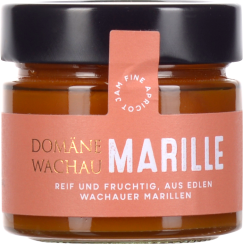 Ein Glas mit dunklem Schraubverschluss, gefüllt mit orangefarbener Marillenmarmelade. Das Etikett ist in Rosa gehalten und hebt sich mit goldener und weißer Schrift ab.