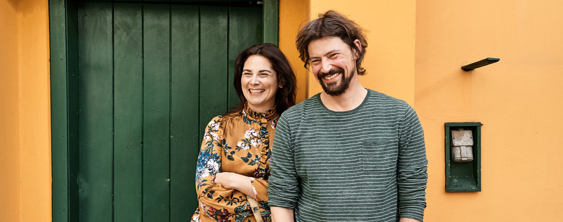 Zwei lächelnde Personen vor gelber Wand mit grüner Tür.