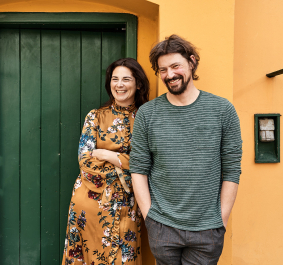 Zwei lächelnde Personen vor gelber Wand mit grüner Tür.
