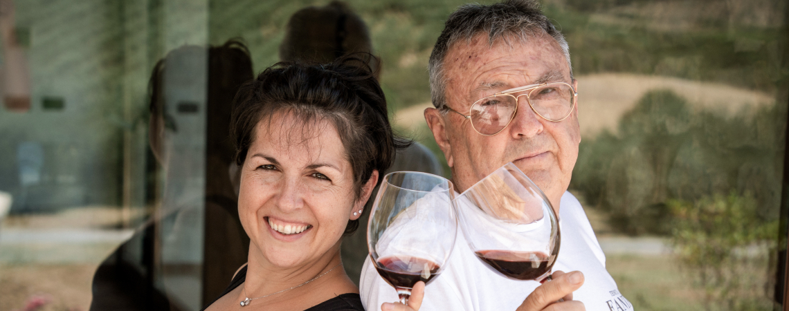 Zwei Personen mit Weingläsern, lächelnd vor einer glasspiegelnden Kulisse.