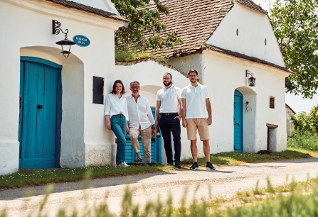 Vier Personen vor weißem Gebäude mit blauen Türen, sonnige ländliche Umgebung.