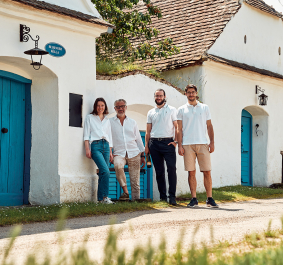 Vier Personen vor weißem Gebäude mit blauen Türen, sonnige ländliche Umgebung.