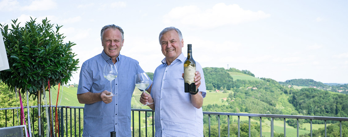 Zwei Männer mit Wein auf Terrasse, malerische Aussicht auf hügelige Landschaft.