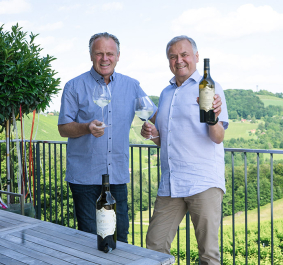 Zwei Männer mit Wein auf Terrasse, malerische Aussicht auf hügelige Landschaft.