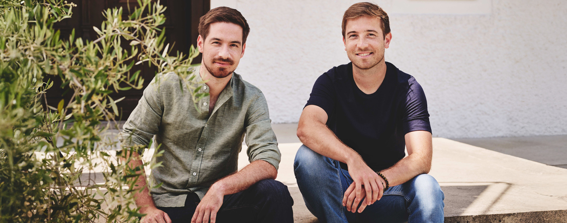 Zwei Männer sitzen auf einer Betonstufe, umgeben von grünen Pflanzen.