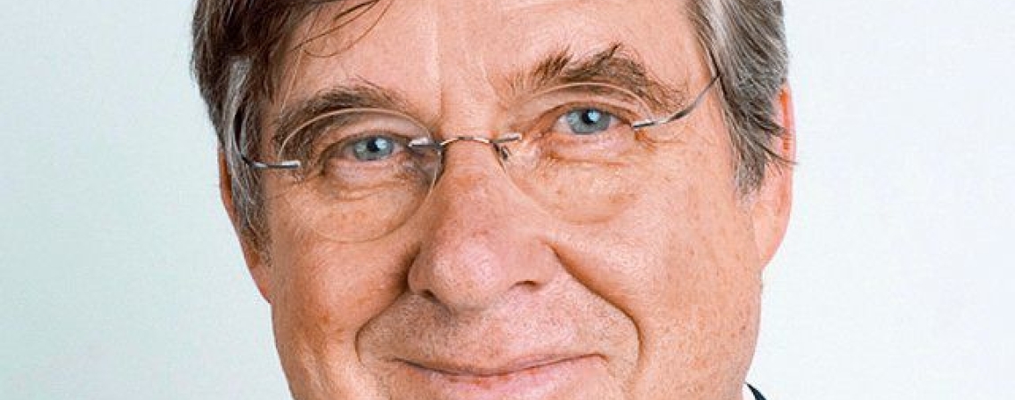 Älterer Mann mit Brille, lächelt, trägt dunklen Anzug und weißes Hemd.