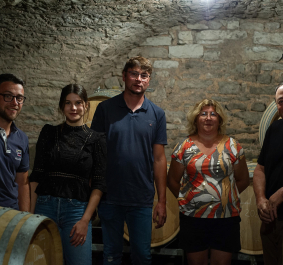 Fünf Menschen in einem Weinkeller mit Holzfässern und gemauerter Steinwand.