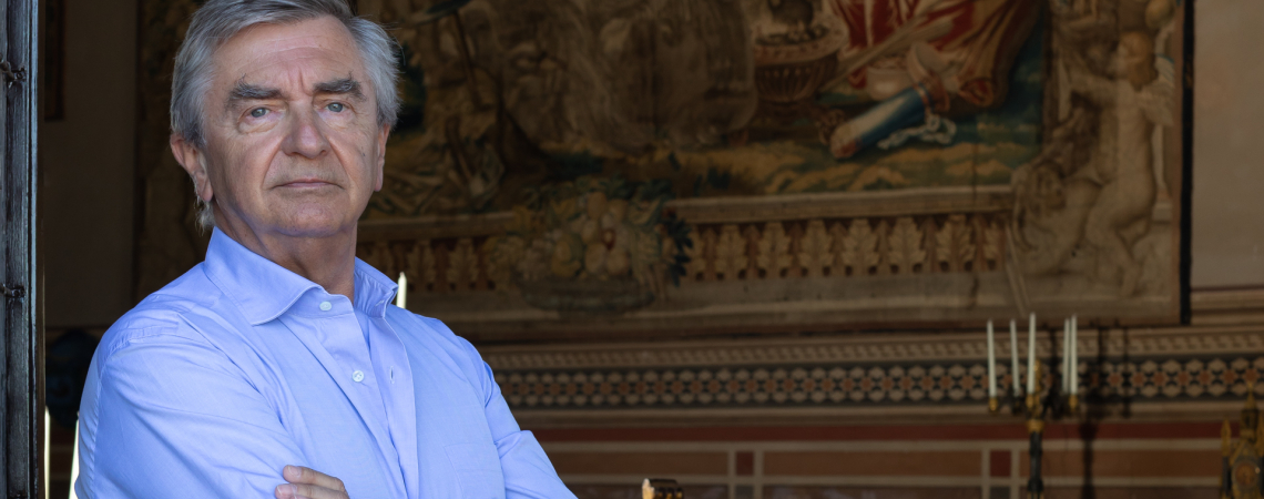 Eleganter Mann in blauem Hemd vor historischer Wandteppichkulisse in edlem Speisesaal.