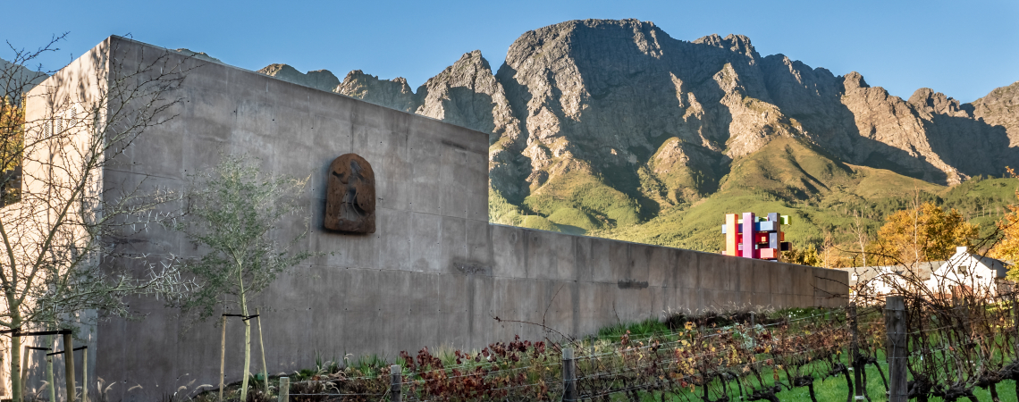 Moderne Betonmauer, Weingarten im Vordergrund, bewaldete Berge im Hintergrund.