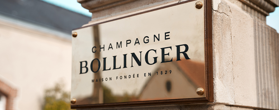 Goldenes Schild an Sandsteinmauer, schwarzer Schriftzug, vor unscharfem Gebäudehintergrund.