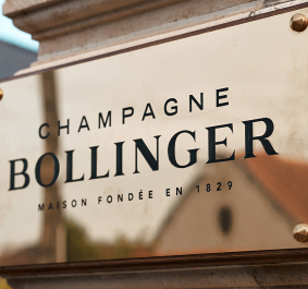 Goldenes Schild an Sandsteinmauer, schwarzer Schriftzug, vor unscharfem Gebäudehintergrund.