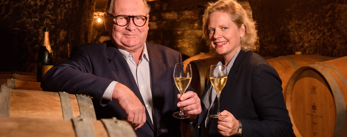 Zwei Personen im Weinkeller stoßen mit Sektgläsern an, umgeben von Holzfässern.