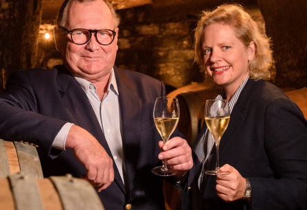 Zwei Personen im Weinkeller stoßen mit Sektgläsern an, umgeben von Holzfässern.