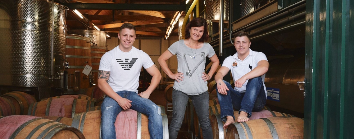 Drei Personen posieren auf Weinfässern in einem Weinkeller.