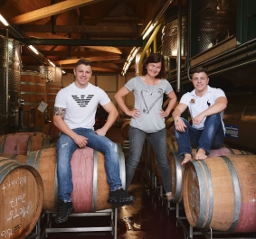 Drei Personen posieren auf Weinfässern in einem Weinkeller.