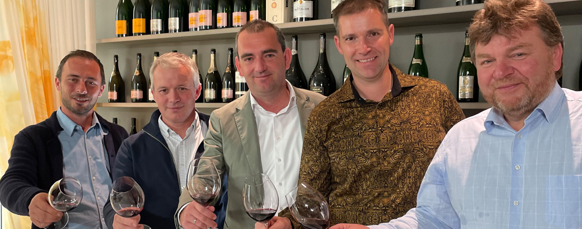 Fünf lächelnde Männer in eleganter Kleidung präsentieren Rotweingläser vor einer Weinregalwand.