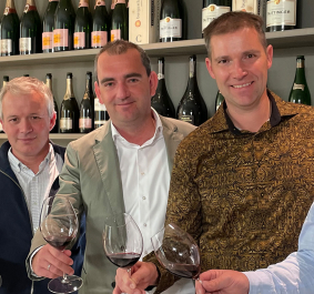 Fünf lächelnde Männer in eleganter Kleidung präsentieren Rotweingläser vor einer Weinregalwand.