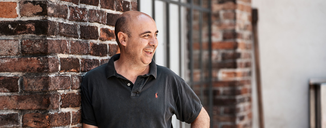 Mann in schwarzem Poloshirt vor Backsteinmauer, lächelnd.