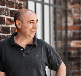 Mann in schwarzem Poloshirt vor Backsteinmauer, lächelnd.