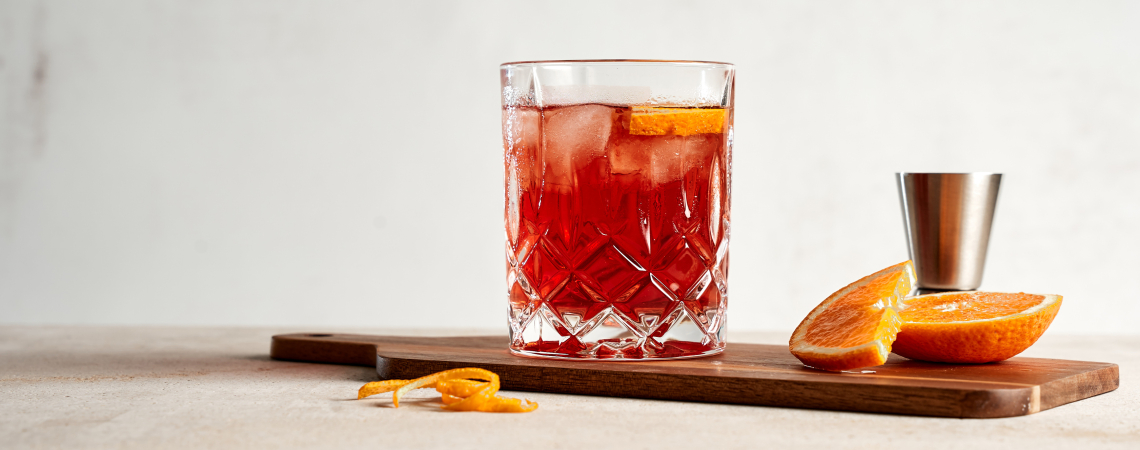 Roter Cocktail im Kristallglas mit Orangenscheibe auf Holzbrett, daneben Orangenstücke.