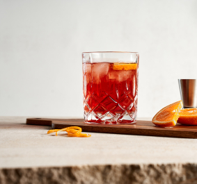 Roter Cocktail im Kristallglas mit Orangenscheibe auf Holzbrett, daneben Orangenstücke.
