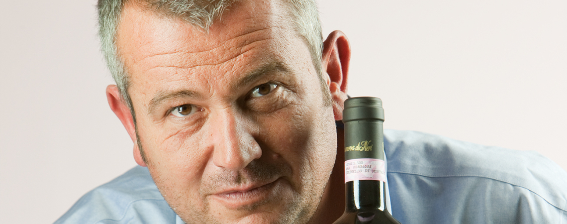 Mann stützt sich auf Holzkiste, hält Weinflasche, trägt blaues Hemd.