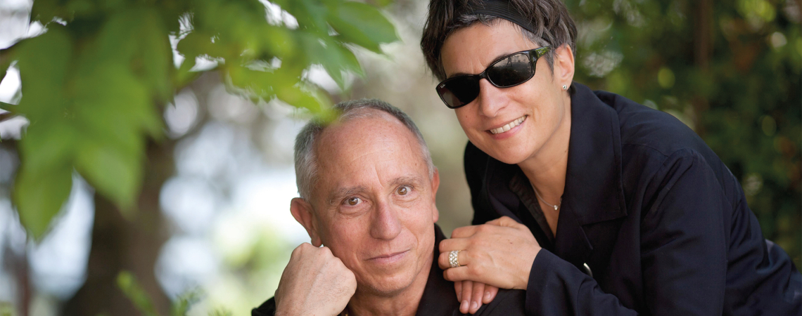 Älteres Paar in schwarzer Kleidung lächelt unter Bäumen, Frau trägt Sonnenbrille.