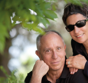 Älteres Paar in schwarzer Kleidung lächelt unter Bäumen, Frau trägt Sonnenbrille.