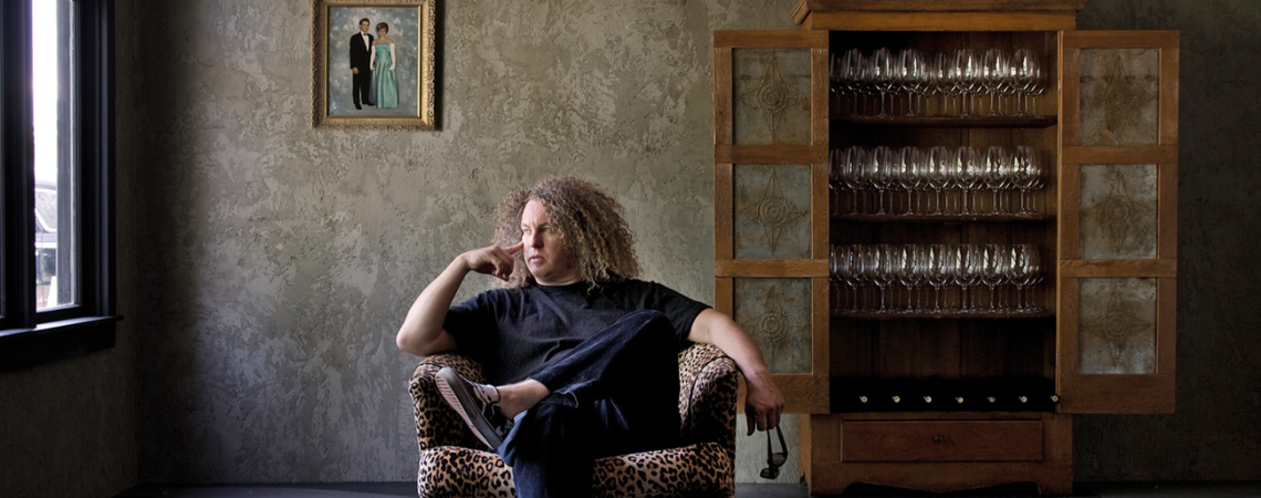 Mann in Leopardenmuster-Sessel, modernes Interieur mit Gläservitrine im Hintergrund.