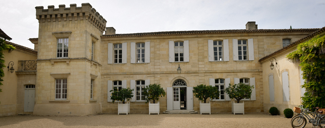 Historisches Steingebäude mit Turm und weißen Fensterläden, umgeben von Pflanzen.