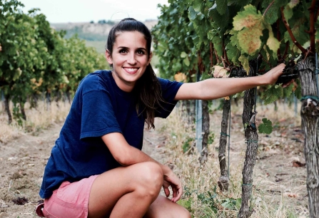 Frau in blauem T-Shirt kniet lächelnd zwischen Weinreben im Weinberg.