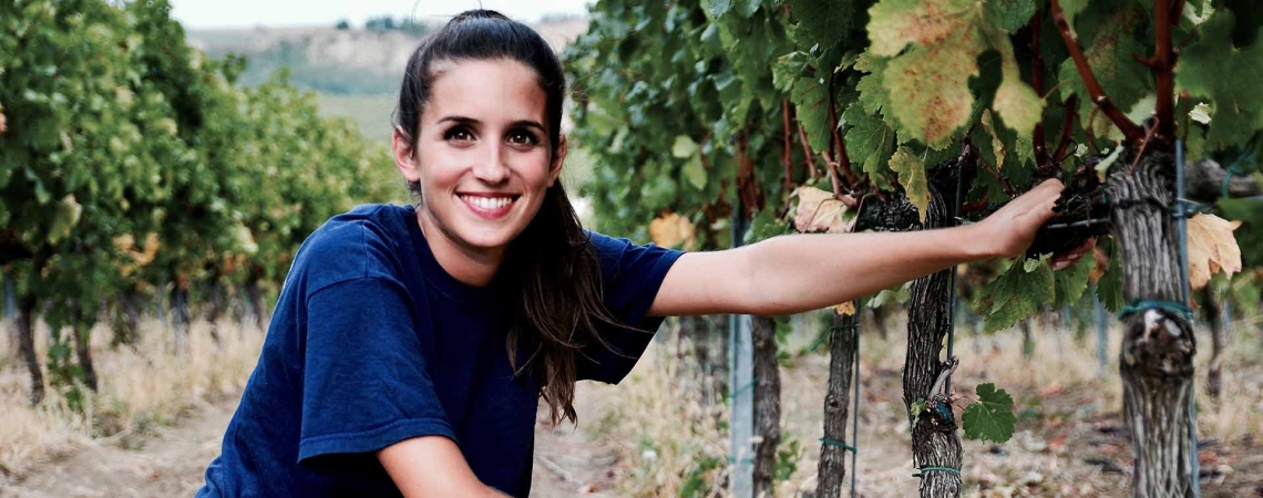 Frau in blauem T-Shirt kniet lächelnd zwischen Weinreben im Weinberg.