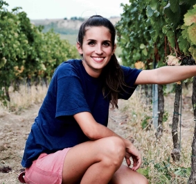 Frau in blauem T-Shirt kniet lächelnd zwischen Weinreben im Weinberg.