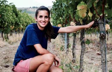Frau in blauem T-Shirt kniet lächelnd zwischen Weinreben im Weinberg.