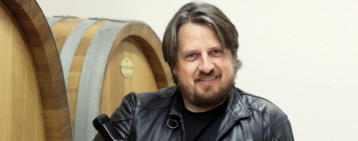 Person in Lederjacke hält große Weinflasche vor hölzernen Weinfässern.