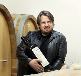 Person in Lederjacke hält große Weinflasche vor hölzernen Weinfässern.