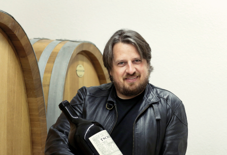 Person in Lederjacke hält große Weinflasche vor hölzernen Weinfässern.